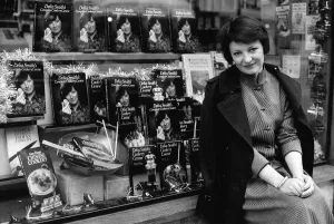 00072 Prints: Delia Smith TV Chef and writer sits outside bookshop that has a window full of her books