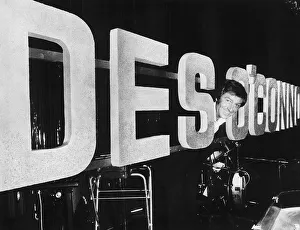 Des O Connor TV Presenter Comedian in fun pose in between enlarged name logo banner