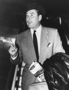 Errol Flynn pointing his finger and holding a suitcase and a news paper with a black