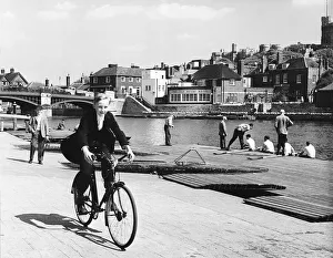 01194 Prints: Eton schoolboy cycling along the Thames in his school uniform Circa 1955