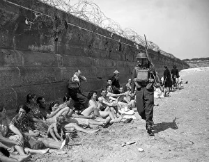 Gas Masks Prints: Holidaymakers sunbathing look pityingly at a passing home front serviceman carrying a
