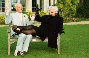 John Inman Comedy Actor and Mollie Sugden Comedy Actress sitting on wooden bench seat in