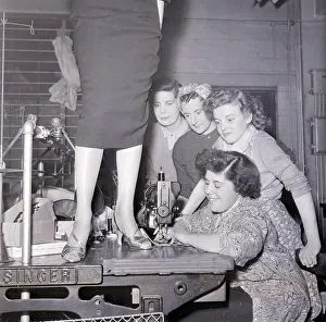 A lady in a pencil skirt stands on a bench while a lady sews on the sewing machine