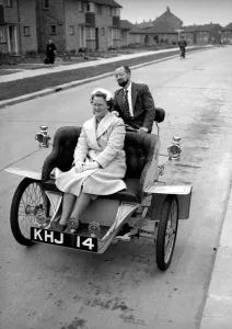 00168 Prints: Laurence Mathew sitting in his veteran car with his wife driving behind May 1956