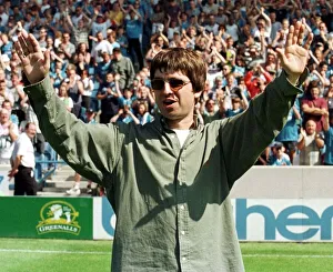 Liam Gallagher arms raised, stands on a pitch at Maine Road during the Premiership match
