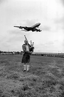 00067 Prints: Lone bagpipe player Jim Thomas playing as a jumbo jet flies overhead May 1975