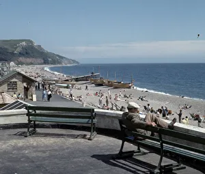 Boats Prints: A man sitting on bench looks out over the beach at the small seaside town of Seaton