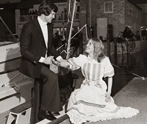 Man wearing a navy blue blazer white polo sweater and trousers helps a lady out of a boat