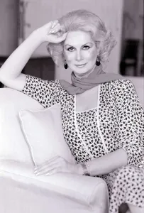 Model April Ashley sitting on a sofa wearing a print dress with one hand at her head May
