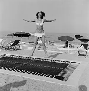 A model wearing bikini swimwear bounces on a trampoline on a beach in the French Riviera