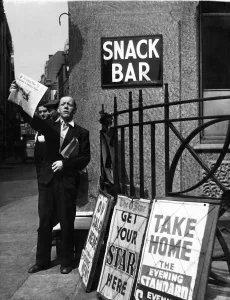 Vendor Prints: A newspaper seller in London, holding a copy of the Evening News newspaper