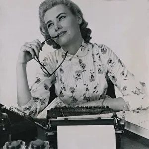 Secretary Prints: Office worker at typewriter chewing the end of her glasses July 1958