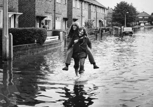 Carrying Prints: Residents of Brunswick Road in Altrincham wade through deep water on the street after