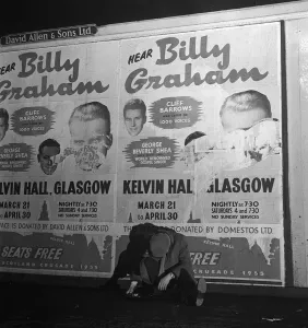 Evangelism Prints: A sleeping drunk sits underneath a defaced poster of Billy Graham in Glasgow ahead of his