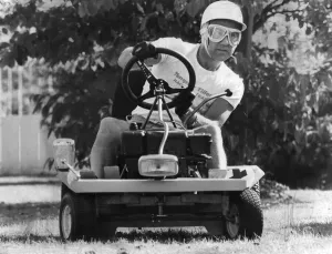 Stirling Moss sitting on lawn mower wearing crash helmet
