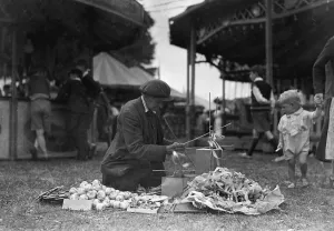 Fairground Prints: A toddler looks on in curiosity as a fancy goods vendor prepares his goods for sale