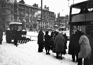 A tractor is used as a snow plough to clear the pavement of snow as people queue for