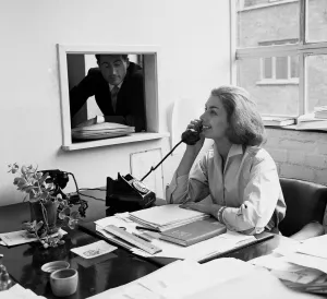 00099 Prints: A woman making a phone call her office desk as a colleague looks through the window