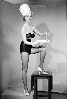 Woman modelling black lingerie and a chefs hat in a cooking photo shoot. Circa 1964
