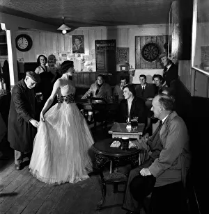 Full Length Prints: Woman showing off a long white dress to a room full of local men during a fashion Show in