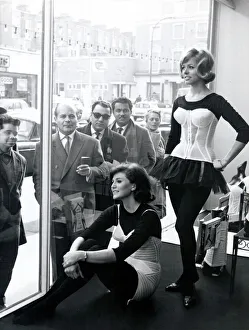 Two women modelling corsets in shop window while people on the street look in - March