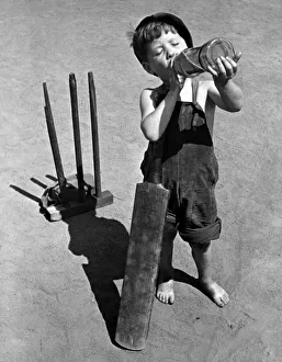 Dungarees Prints: Five year old boy enjoys a refreshing drink from a bottle whilst playing cricket on a hot