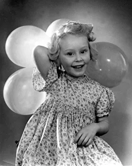 Birthdays Prints: Young girl holding balloons for a party wearing floral patterned dress Circa 1945
